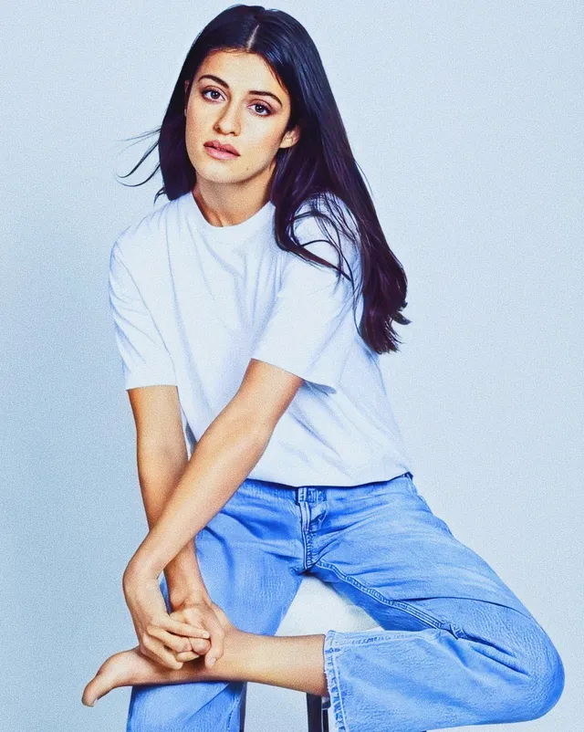 Anya Chalotra sitting barefoot in blue jeans and a white shirt.