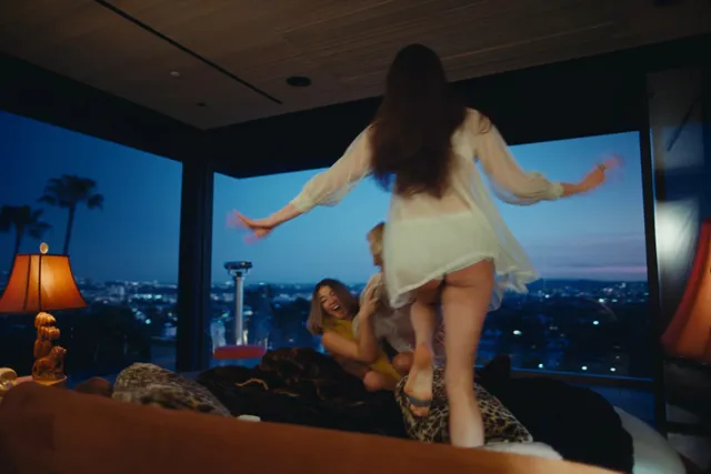 Margaret Qualley a screen scene dancing on a sofa in a white outfit.
