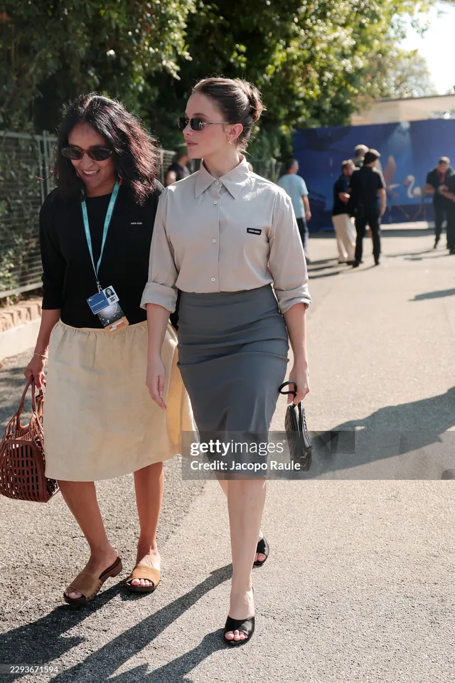 Cailee Spaeny walking outdoors in a pale shirt, grey skirt and heels.