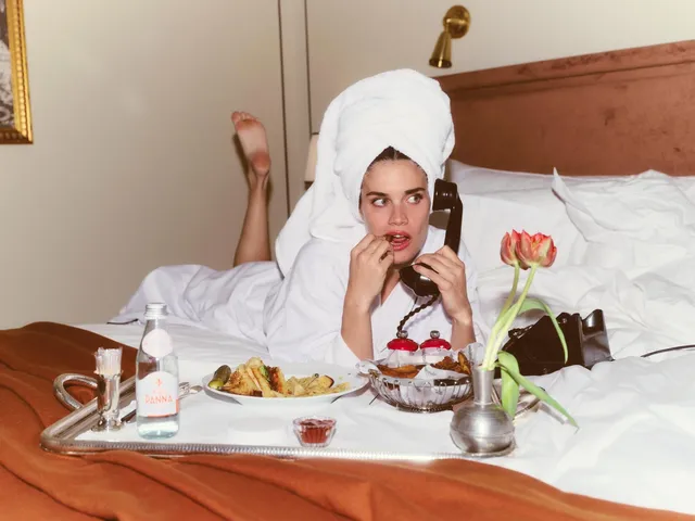 Sara Sampaio reclining on a bed in a white robe and heeled sandals with a breakfast tray.