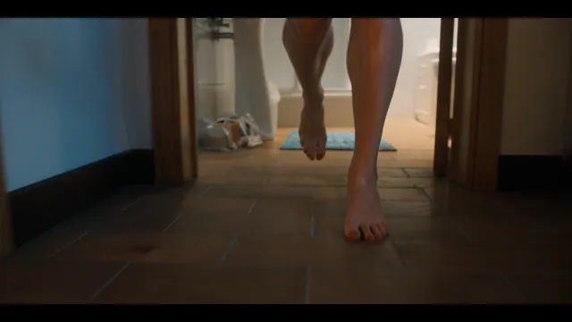 Grace Van Patten raising one bare foot while walking indoors in a film scene.