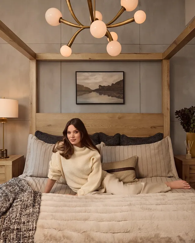 Hailee Steinfeld sitting barefoot on a bed in a pale outfit.