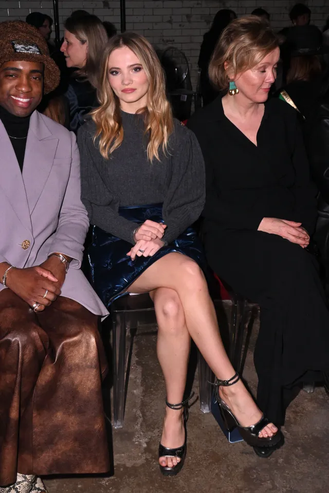 Freya Allan sitting beside two people at an event in black heels.