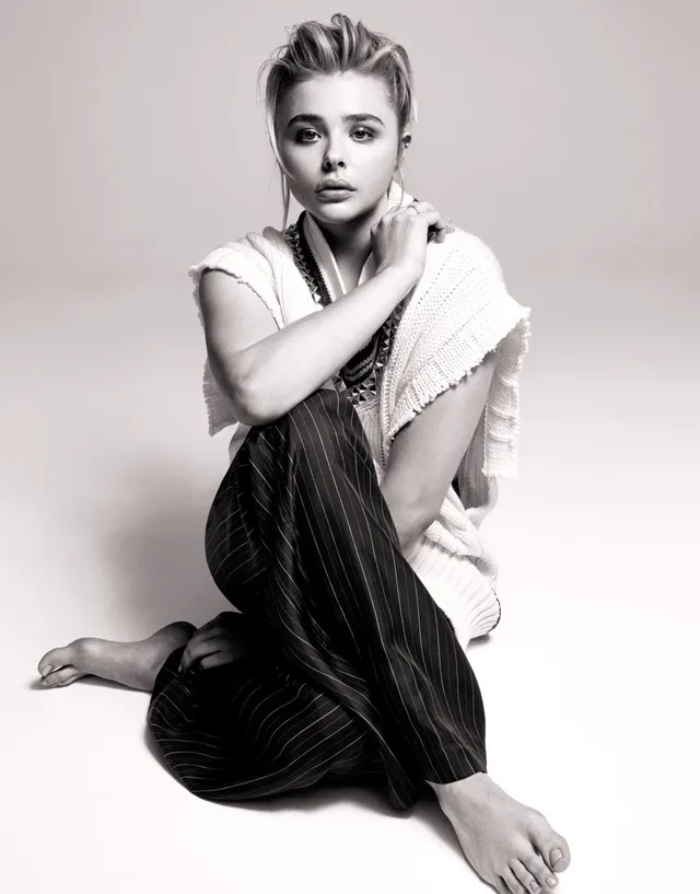 Chloë Grace Moretz sitting barefoot in a monochrome studio portrait.