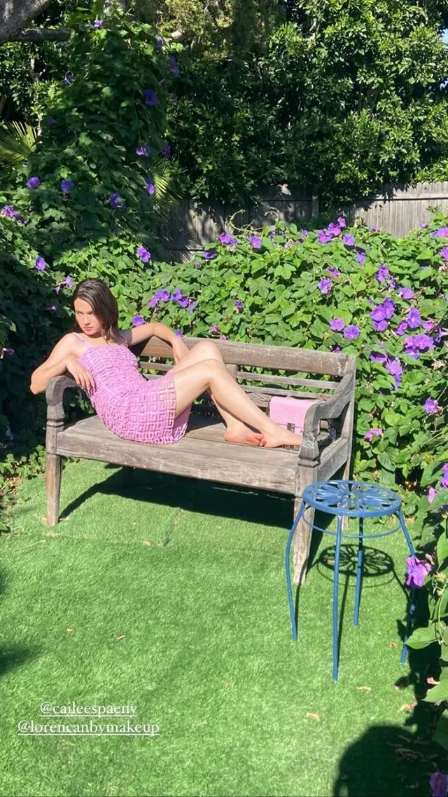 Cailee Spaeny relaxing barefoot on a wooden bench in a flowering garden.