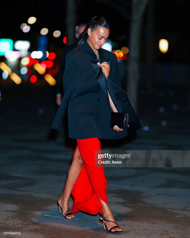 Zoe Saldaña walking outdoors in a black jacket and red skirt.