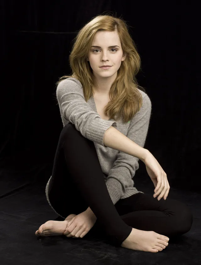 Emma Watson: seated barefoot in black trousers against a dark background.