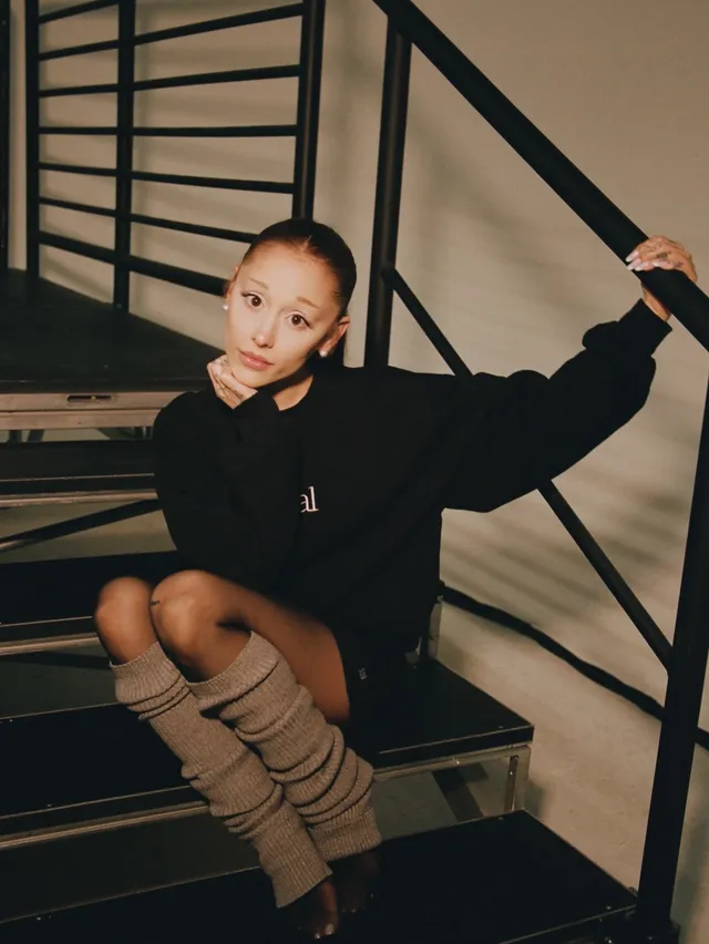 Ariana Grande: seated on stairs in a black outfit and tall high heels.
