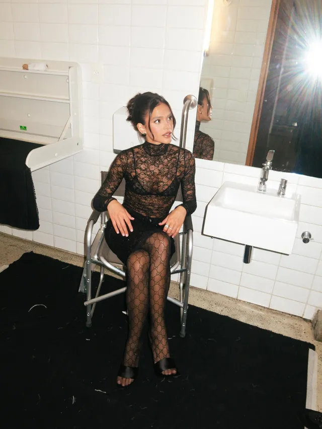 Bruna Marquezine sitting in a dressing room in a black lace outfit and heels.