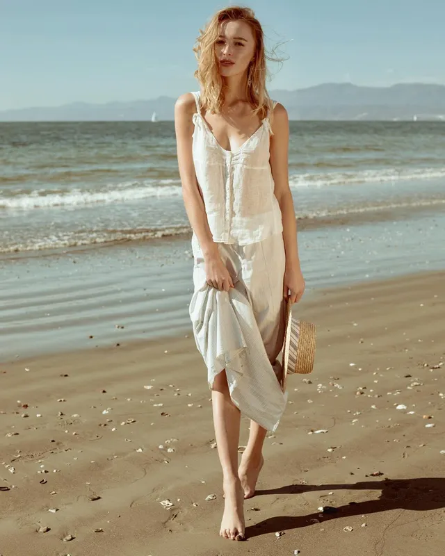 Phoebe Dynevor standing barefoot on a sandy beach.