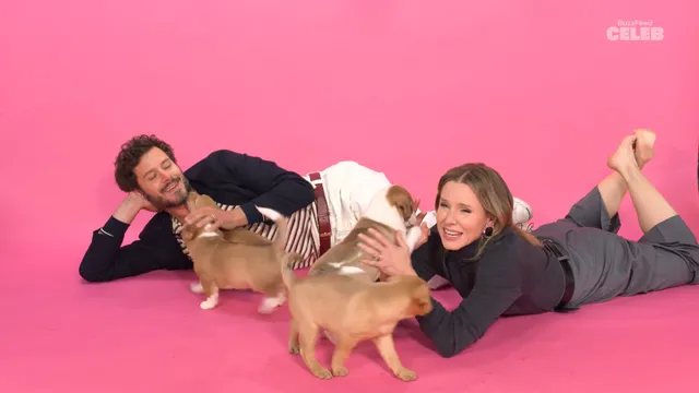 Kristen Bell lying with bare feet raised beside small dogs on a pink backdrop.