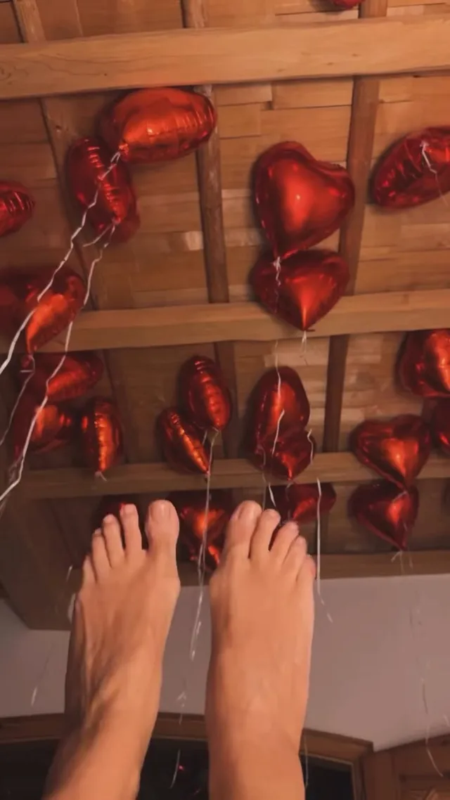 Heidi Klum standing barefoot beside red heart-shaped balloons on a wooden floor.