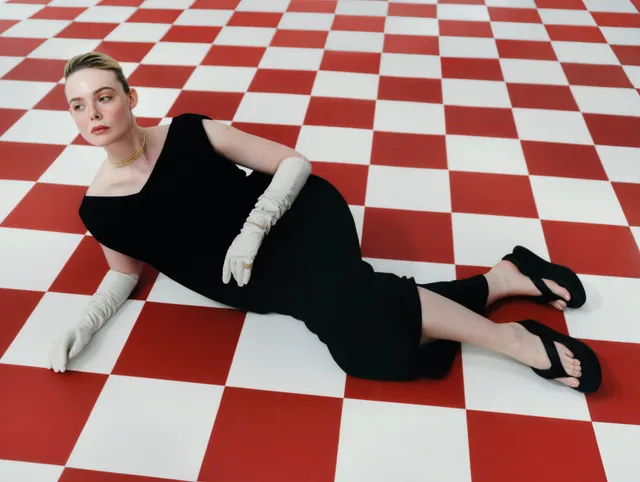 Elle Fanning posing on a red-and-white tiled floor in a black dress and heels.
