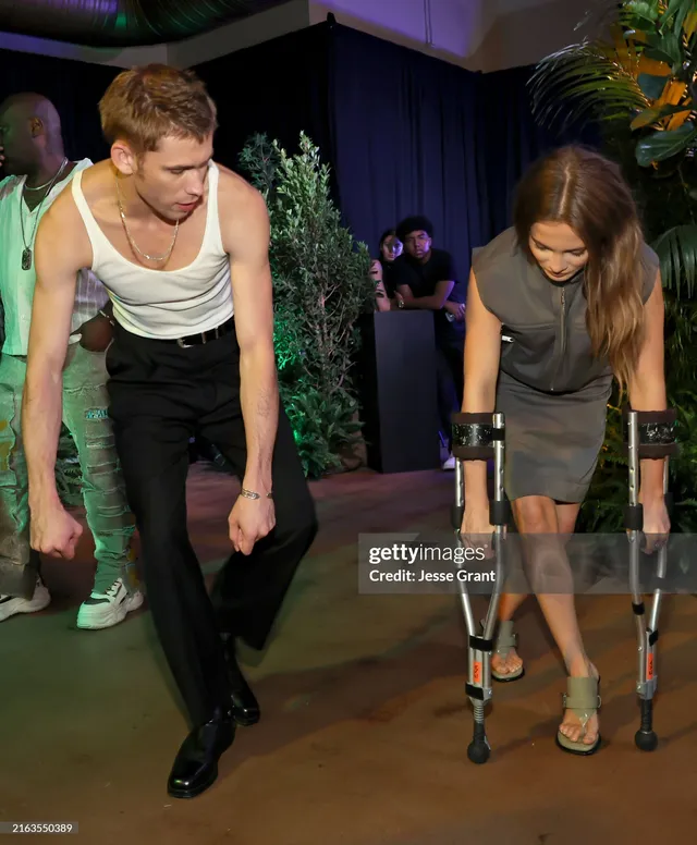 Freya Allan leaning over a table beside a man in metallic heels.