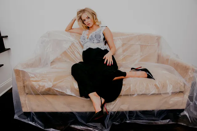 Samara Weaving reclining on a sofa in a dark outfit and black heels.