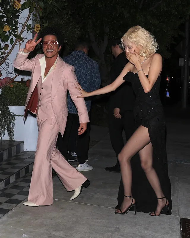 Rosé walking beside a guest in a pink suit in a black dress and strappy heels.