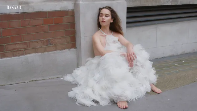 Emilia Clarke sitting barefoot on a pavement in a feathered dress.