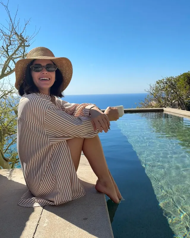Lucy Hale sitting on the edge of an infinity pool in a striped outfit.