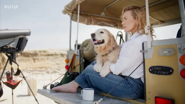 Kate Winslet sitting barefoot in a vehicle beside a dog.