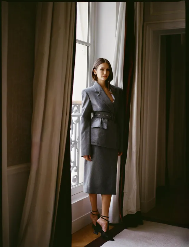 Nina Dobrev wearing a gray coat and black heels by a window.