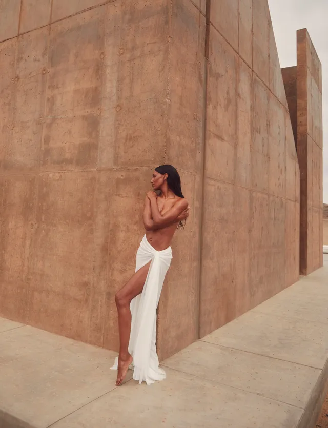 Jasmine Tookes standing barefoot against a stone wall wearing a pale skirt.