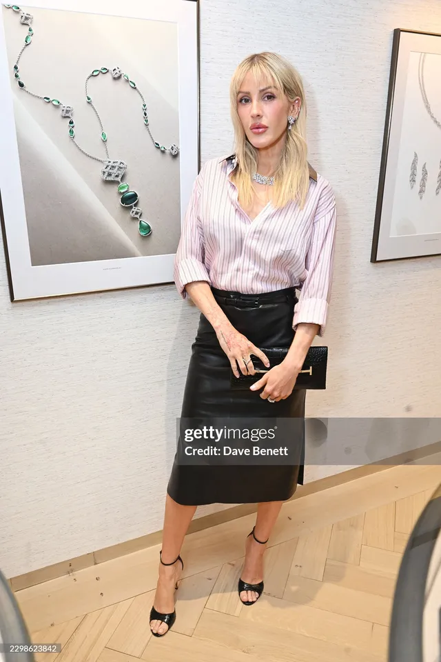 Ellie Goulding standing indoors in a pale blouse, black skirt and heels.