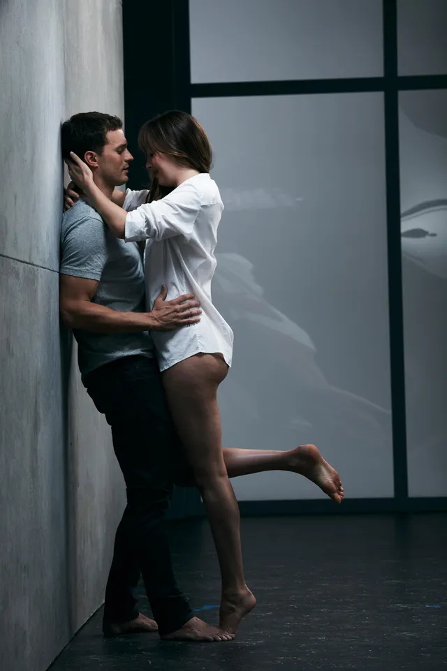 Dakota Johnson standing barefoot with a companion by a window.