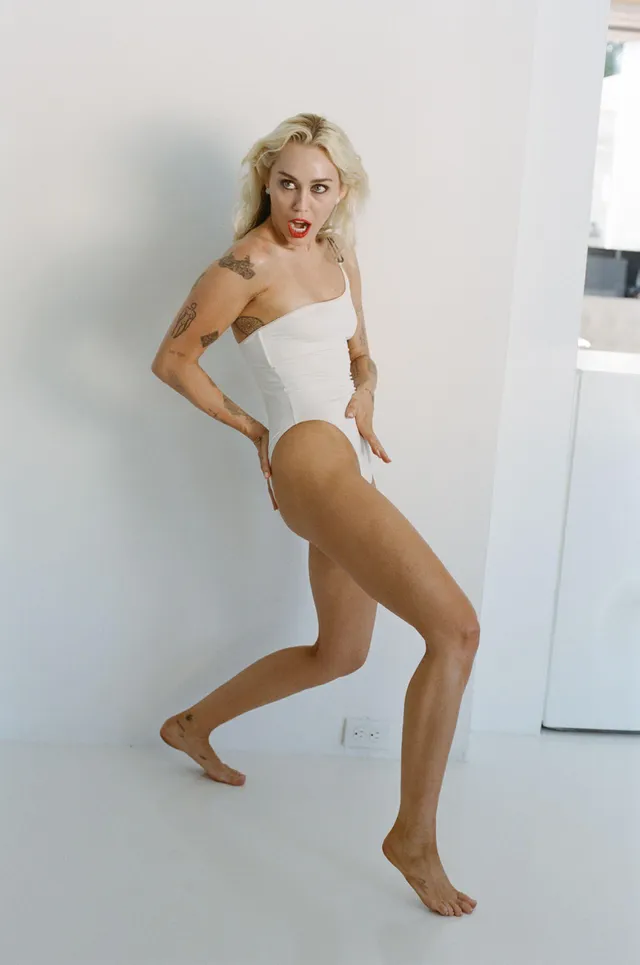 Miley Cyrus standing barefoot in a white studio wearing a pale bodysuit.