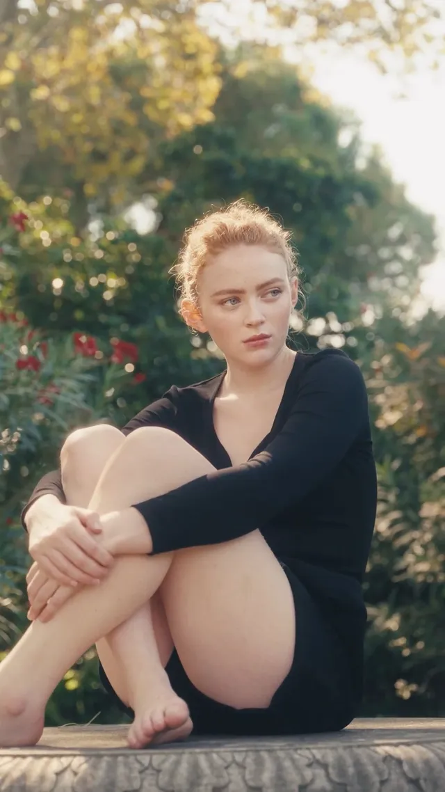 Sadie Sink sitting barefoot outdoors in a black outfit.