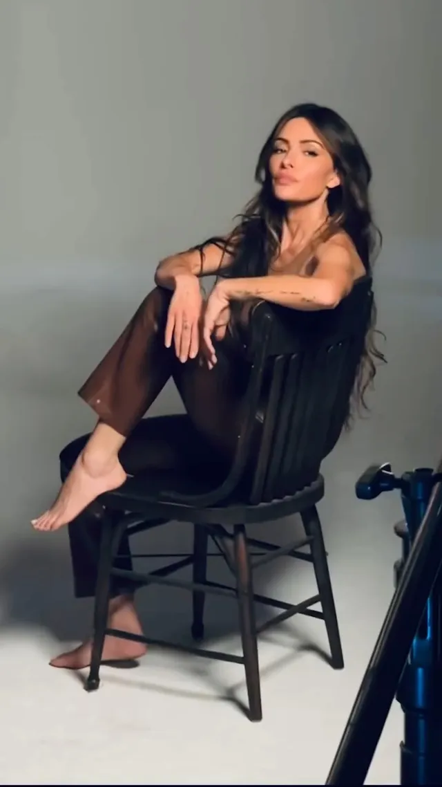 Sarah Shahi sitting in a wooden chair in a dark outfit.