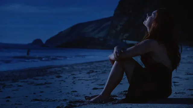 Mackenzie Ziegler sitting barefoot on beach sand in a dark scene.