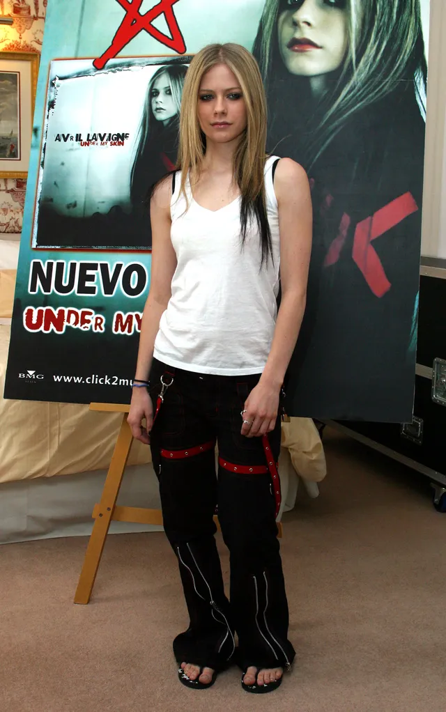 Avril Lavigne standing indoors in dark trousers and flip-flops.