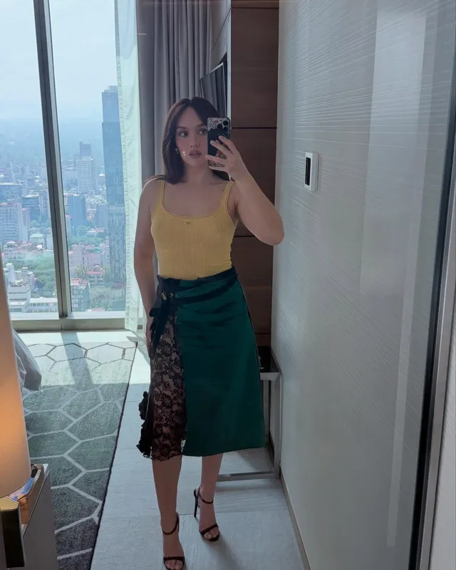 Olivia Cooke taking a mirror photograph in a yellow top and green skirt.