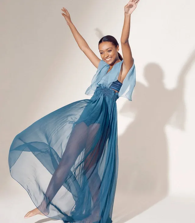 Sophie Wilde jumping barefoot in a blue gown with her arms raised.