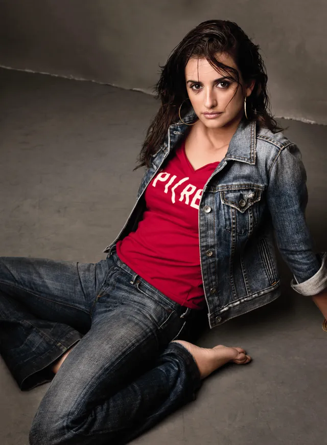 Penélope Cruz sitting barefoot in jeans and a denim jacket for a portrait.