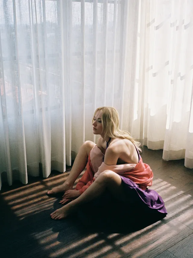 Maika Monroe sitting barefoot by a curtain.