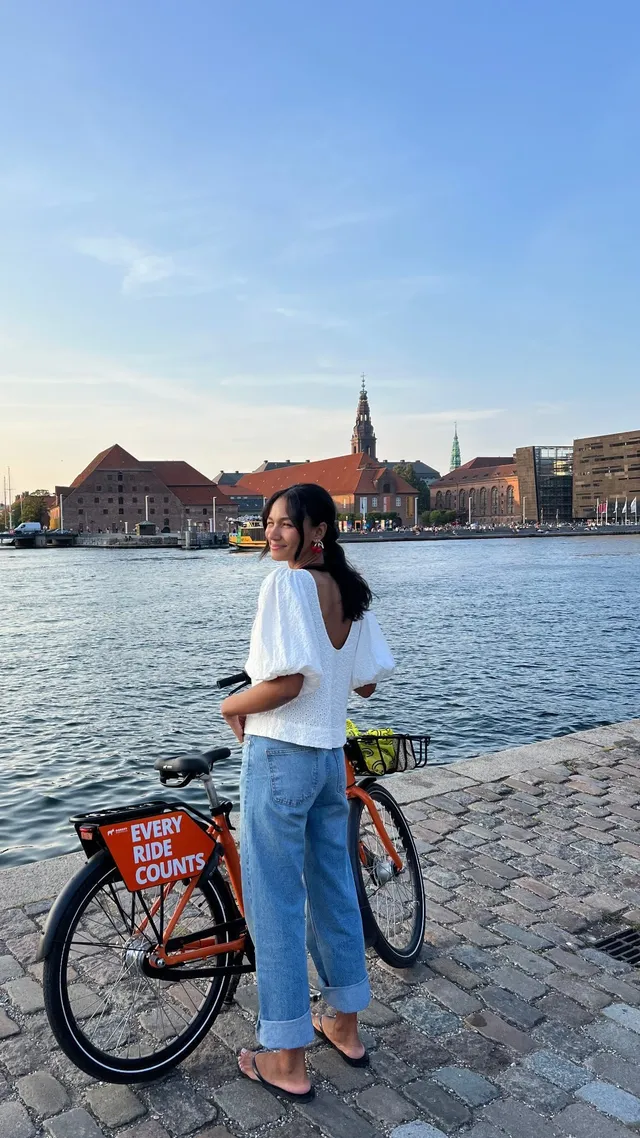 Greta Onieogou standing beside a bicycle in a white top, jeans and flats.