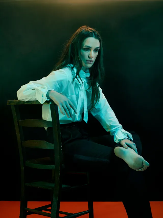 Keira Knightley seated in a dark chair with one bare sole raised.