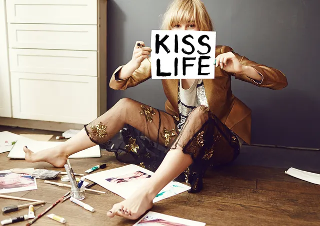 Suki Waterhouse sitting on a floor holding a sign.