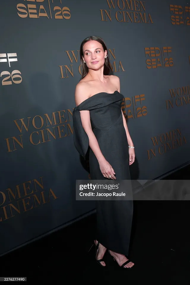 Alicia Vikander posing against a dark event backdrop in a black dress.