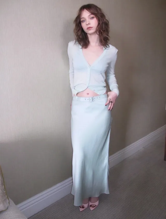 Emma Myers posing in a pale skirt suit and pink heels indoors.