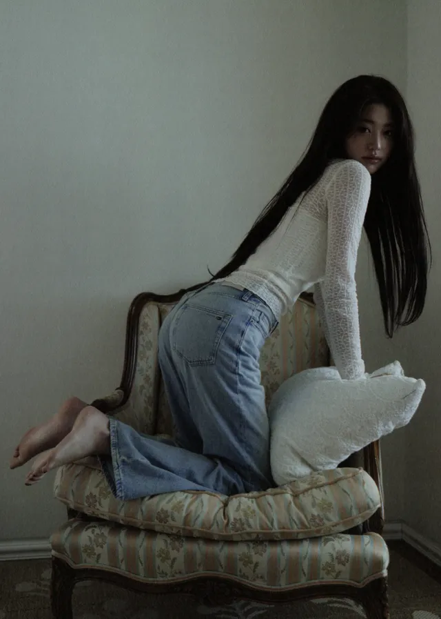 Jang Wonyoung kneeling barefoot on an upholstered chair in jeans.