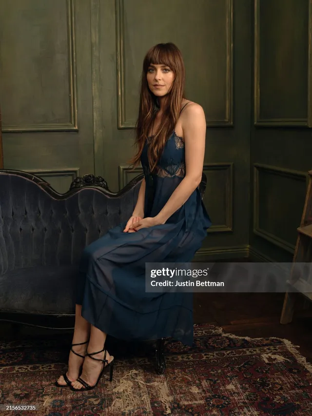 Dakota Johnson sitting in a dark dress and strappy heels.