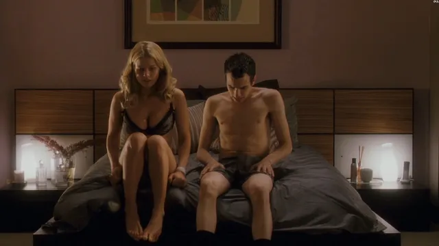 Alice Eve talking with another character while seated on a bed.