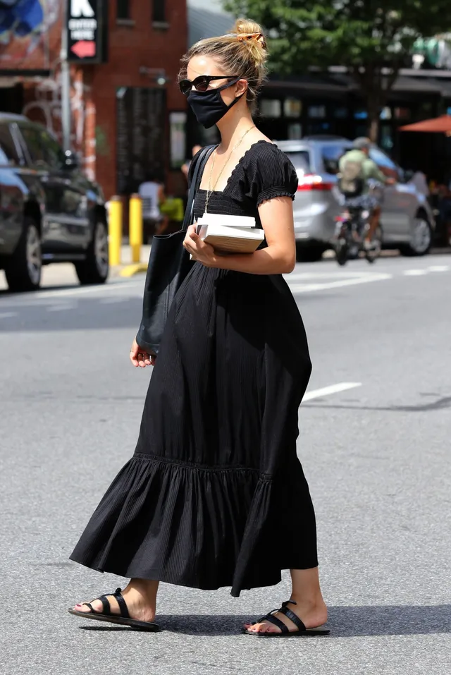 Dianna Agron walking beside a vehicle in a black dress and flat sandals.