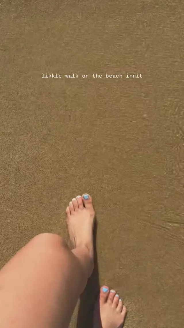 Rachel Zegler walking barefoot over wet sand with painted toenails.