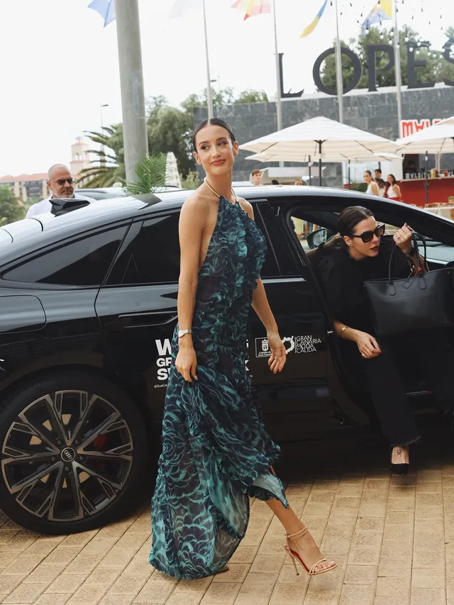 María Pedraza walking beside a vehicle in a patterned gown and heels.