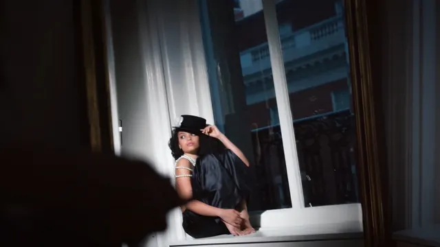 Whitney Peak sitting barefoot on a window ledge in a dark outfit and hat.