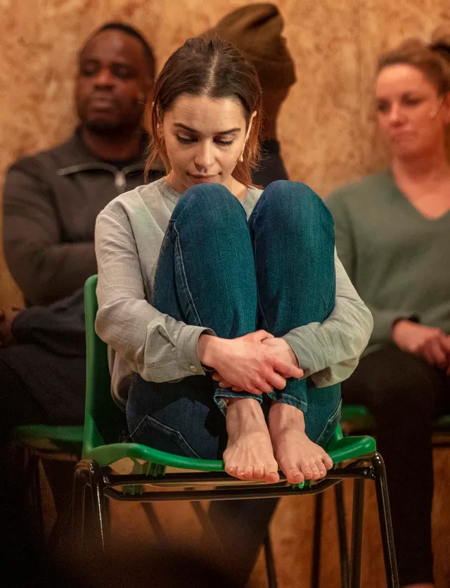 Emilia Clarke sitting barefoot on a green chair in jeans.
