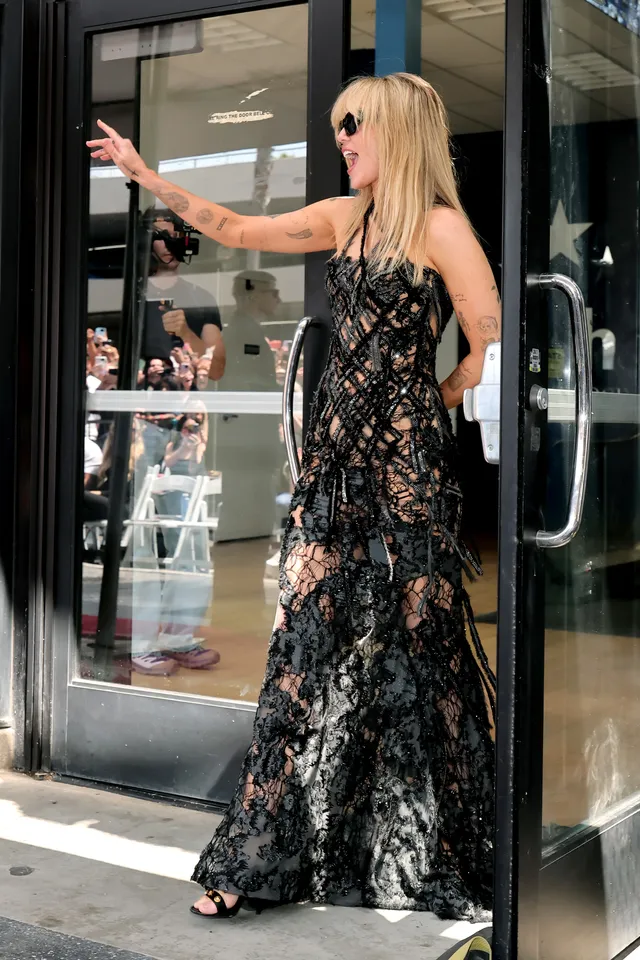 Miley Cyrus standing in a black dress and heels outdoors.